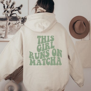Könnte beinhalten: Ein cremefarbener Kapuzenpullover mit dem Text THIS GIRL RUNS ON MATCHA, der in einer großen, wellenförmigen, salbeigrünen Schrift auf dem Rücken gedruckt ist. Das Kleidungsstück hat lange Ärmel, eine Kapuze und eine lockere Passform.