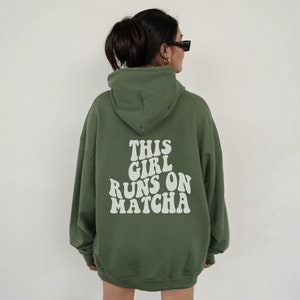 Könnte beinhalten: Ein olivgrüner Kapuzenpullover mit dem weißen Schriftzug THIS GIRL RUNS ON MATCHA auf der Rückseite in einer fetten, wellenförmigen Schrift. Das Kleidungsstück verfügt über eine Kapuze, lange Ärmel mit gerippten Bündchen und eine lockere Passform.