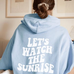 May include: A light blue hoodie with the text "Let's Watch The Sunrise" in white retro-style lettering.