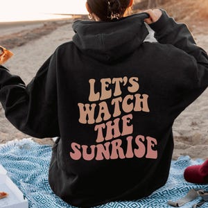 May include: A black hoodie with the text "Let's watch the sunrise" in pink and orange letters. The person wearing the hoodie is holding a slice of pizza.