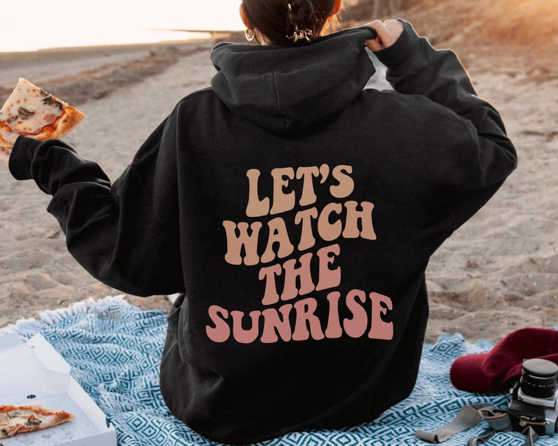 May include: A black hoodie with the text "Let's watch the sunrise" in pink and orange letters. The person wearing the hoodie is holding a slice of pizza.