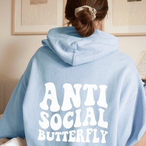 May include: Light blue hoodie with the words "ANTI SOCIAL BUTTERFLY" in white, retro-style font across the back. The hoodie has a hood and long sleeves. The image is a close-up of the back of the hoodie.