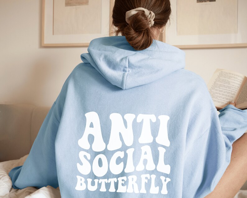 May include: Light blue hoodie with the words "ANTI SOCIAL BUTTERFLY" in white, retro-style font across the back. The hoodie has a hood and long sleeves. The image is a close-up of the back of the hoodie.