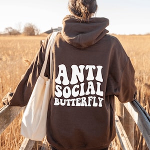 May include: A brown hoodie with the words "ANTI SOCIAL BUTTERFLY" in white, retro-style font. The hoodie is worn by a person standing on a wooden bridge, with a beige tote bag over their shoulder. The background features tall, dry grass.