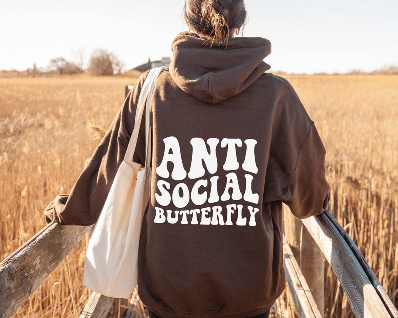 May include: A brown hoodie with the words "ANTI SOCIAL BUTTERFLY" in white, retro-style font. The hoodie is worn by a person standing on a wooden bridge, with a beige tote bag over their shoulder. The background features tall, dry grass.