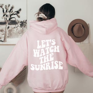 May include: A pink hoodie with a white graphic that says "Let's Watch The Sunrise" in a retro font.