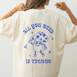 Puede incluir: Camiseta color crema con un dise&ntilde;o de hongo azul de dibujos animados y el texto "ALL YOU NEED IS TECHNO". El hongo tiene una cara sonriente y hace el signo de la paz. El dise&ntilde;o incluye notas musicales y estrellas.