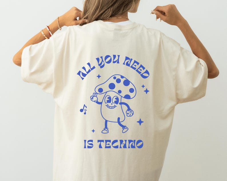 Puede incluir: Camiseta color crema con un dise&ntilde;o de hongo azul de dibujos animados y el texto "ALL YOU NEED IS TECHNO". El hongo tiene una cara sonriente y hace el signo de la paz. El dise&ntilde;o incluye notas musicales y estrellas.
