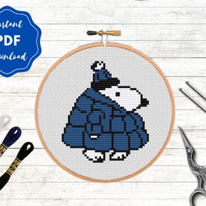 May include: A cross-stitch embroidery hoop featuring a cartoon character in a blue coat and hat. The design is set against a white background with a wooden hoop. The image includes embroidery floss, needles, and scissors. Text reads "Instant PDF Download."