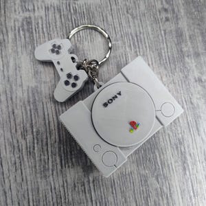 May include: A white PlayStation 1 keychain with a grey controller attached. The keychain has the word "SONY" printed on it.