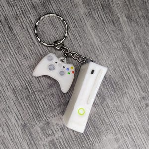 May include: White plastic keychain featuring a miniature Xbox console and controller. The console has a green power button and the controller has coloured buttons.