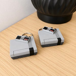 May include: Two grey and dark grey keychains shaped like retro video game consoles, each featuring a small attached controller charm with a black directional pad and two red buttons, connected by a silver metal ring for keys.