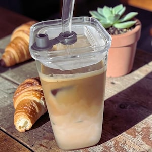 Op de afbeelding: Een doorzichtige plastic beker met ijskoffie, een deksel en een rietje, gevuld met een gelaagde koffiedrank. Twee croissants liggen op een houten oppervlak naast de beker. Een kleine vetplant staat op de achtergrond.