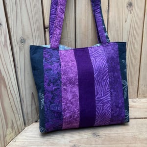 Peut inclure: Un sac fourre-tout patchwork avec une variété de tissus violets. Le sac présente un motif rayé avec différents motifs et nuances de violet, y compris des imprimés floraux et abstraits. Les poignées sont violettes unies. Le sac est posé sur une surface en bois.