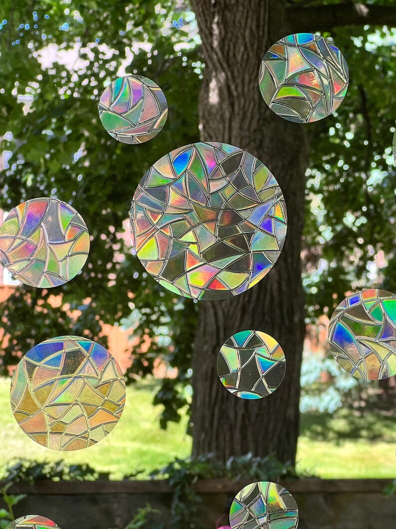 May include: A window with a view of a tree and green grass. The window is decorated with iridescent, circular stickers that resemble stained glass.