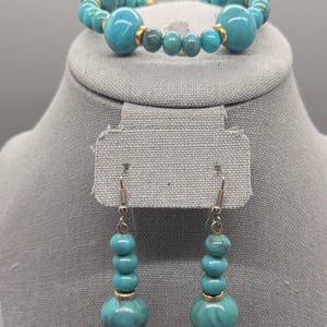 May include: A turquoise beaded bracelet and matching earrings set. The bracelet features large, round beads with gold-coloured accents. The earrings have a similar design, with multiple turquoise beads suspended from silver-coloured hooks. The jewellery is displayed on a grey surface.