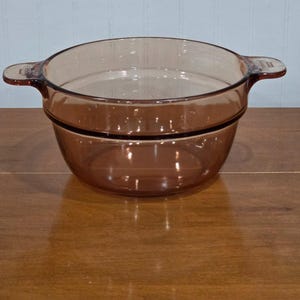 May include: A vintage amber-colored glass casserole dish with two handles. The dish is round with a slightly flared rim and a dark band around the middle. It sits on a wooden surface.