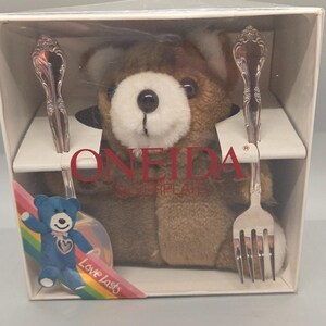 Peut inclure: Un ours en peluche marron emballé dans une boîte avec une cuillère et une fourchette en métal argenté. La boîte porte le texte ONEIDA SILVERPLATE et un petit graphique d'un ours bleu avec un arc-en-ciel et le texte Love Lasts en bas à gauche.