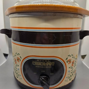 May include: Vintage Rival Crock-Pot slow cooker with a clear glass lid and brown rim. The cream-colored pot has a black band with orange and blue stripes, floral accents, and the text "RIVAL CROCK-POT". It has a removable server and a dial for temperature control.