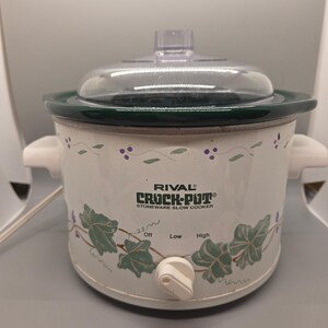 May include: A white Rival Crock-Pot stoneware slow cooker with a clear glass lid and dark green rim. The exterior features a green ivy leaf and purple berry vine pattern. It has white side handles and a front control knob with Off, Low, and High settings.