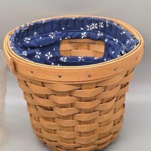 Può includere: Un cesto rotondo in legno intrecciato con una finitura naturale marrone chiaro e una fodera in tessuto blu scuro. La fodera è decorata con un motivo floreale bianco ripetuto ed è arricciata sul bordo superiore, fissato con piccoli chiodi metallici.