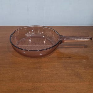 May include: A clear, amber-coloured glass skillet with a long handle. The skillet is round with a slightly flared rim and is sitting on a wooden surface. The handle extends horizontally from the side of the skillet.