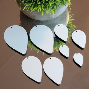 May include: Seven blank, white, teardrop-shaped wooden ornaments with holes for hanging. The ornaments are arranged in a semi-circle on a reflective surface.