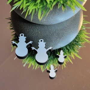 May include: Four white, cutout, snowman ornaments with black hats. The ornaments are on a reflective surface with green grass in the background.