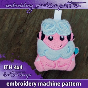 Puede incluir: Juguete de peluche bordado en forma de criatura rosa y azul sobre fondo blanco. El juguete está diseñado para ser utilizado con una máquina de bordar y está etiquetado como "ITH 4x4 in the hoop" y "embroidery machine pattern".
