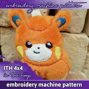 May include: An orange plush toy in the shape of a cartoon animal with blue and yellow accents. The text "embroidery machine pattern" and "ITH 4x4 in the hoop" are visible. The toy has a smiling face and is made of soft, fuzzy material.