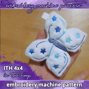 Pode incluir: Uma borboleta de pelúcia branca com estrelas bordadas azuis e um contorno bordado azul. A borboleta mede 4x4 polegadas e é um padrão de máquina de bordado no aro.