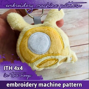 Könnte beinhalten: Ein gelbes Plüschtier mit einem weißen Kreis in der Mitte. Das Spielzeug ist an einem silbernen Schlüsselring befestigt. Das Spielzeug ist mit "ITH 4x4 in the hoop embroidery machine pattern" beschriftet.