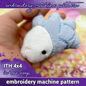 Puede incluir: Un juguete de peluche azul y blanco con forma de pez. El juguete está siendo sostenido en una mano. El texto "ITH 4x4 in the hoop embroidery machine pattern" es visible en la imagen.