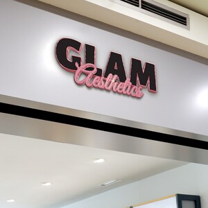May include: A shopfront sign with the text "GLAM Aesthetics" in black and pink 3D lettering.