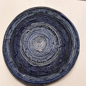 Puede incluir: Un cuenco redondo de mezclilla hecho a mano. El cuenco está construido con tiras de tela vaquera azul enrolladas, creando un patrón en espiral. Los tonos de mezclilla varían de azul claro a oscuro, con algunos hilos blancos visibles. Diámetro: aprox. 20 cm.
