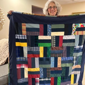 May include: A patchwork quilt with a navy blue border. The quilt features rectangular patches in various colors and patterns, including plaid, floral, and solid fabrics. The quilt is held up, showcasing its size and design.