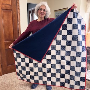 Könnte beinhalten: Eine handgefertigte Steppdecke mit marineblauem Rücken und einem Schachbrettmuster aus weißen Quadraten und marineblauen Blumenquadraten. Die Steppdecke ist mit einem roten Rand versehen.