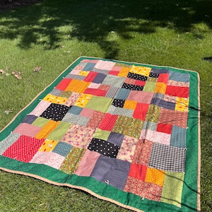 May include: A rectangular patchwork quilt featuring a variety of fabric squares in red, yellow, green, blue, black, and white with floral, polka dot, and geometric patterns. The quilt is finished with a solid dark green border and tan trim.