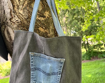 May include: A dark grey tote bag with light blue denim handles and a denim pocket. The bag is hanging from a black metal hook on a tree. The background is blurred green foliage and grass.