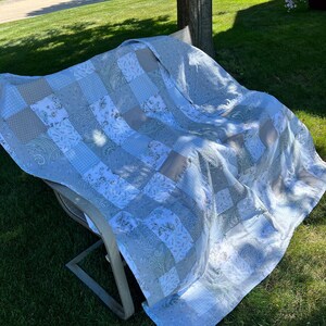 Puede incluir: Un edredón patchwork sobre una silla al aire libre. El edredón presenta cuadrados con diseños florales y de cachemira en tonos azul claro, blanco y gris. El edredón de tela suave se exhibe en un día soleado.