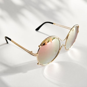 May include: Round sunglasses featuring a thin gold-tone metal frame and reflective pink-tinted lenses. The temples are slender with black plastic tips at the ends. The design includes a double-rimmed circular lens structure with a bridge connecting them.