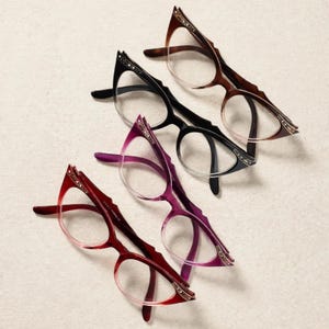 May include: Four pairs of cat-eye glasses arranged diagonally. The frames feature a gradient color transition from dark to light, including brown, black, purple, and red. Each temple arm is decorated with small, embedded metallic studs along the top edge.