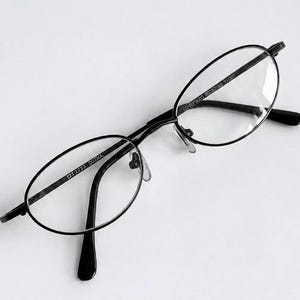 Pode incluir: Óculos de formato oval com uma armação fina de metal preto e lentes transparentes. O interior da haste esquerda apresenta o texto 1733 CHINA, enquanto a lente direita tem um texto ténue impresso. Inclui almofadas de nariz ajustáveis.