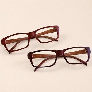 May include: Two pairs of rectangular eyeglasses with dark reddish-brown wood grain frames. The inner temples of the frames feature the text "HAND MADE FRAME" printed in a light color. The glasses have clear lenses and a smooth, polished finish.