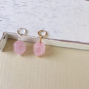 May include: Two rose quartz gemstone charms. One charm has a silver-colored loop, and the other has a gold-colored loop. Both charms have a small silver or gold bead at the bottom. The gemstones are a light pink color and are hexagonal in shape.