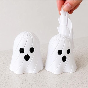 Pode incluir: Duas figuras brancas em forma de fantasma com olhos e bocas circulares pretos. As figuras apresentam um design em espiral com camadas e detalhes de costura visíveis no topo. Uma parte superior é mostrada sendo levantada para cima.