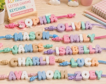 May include: A collection of chunky alphabet letter charms on colorful cords, forming names like Sophia, Olivia, Mason, Emily, James, Isabella, Charlie, Luca, Ava, Noah, and Zoey. The charms are in pastel shades, and a sign reads 3D CHUNKY ALPHABET LETTER CHARMS KEYCHAIN.