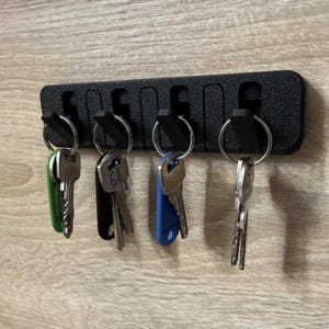 May include: A black rectangular wall-mounted key holder with a textured finish featuring six vertical slots and four angled hooks. Four metal keys are attached to the hooks, including keys with green, black, and blue plastic covers on their heads.