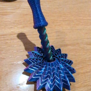May include: A 3D printed spinning top featuring a central twisted stem and a wide, multi-layered base with a jagged, star-like edge. The object displays a gradient of iridescent blue, purple, and green colours with a visible textured finish.