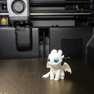 May include: A small, white, 3D-printed dragon figurine with large, bright blue eyes and small wings, sitting on the textured black build plate of a Bambu Lab 3D printer. The printer's print head is visible in the background above the figurine.